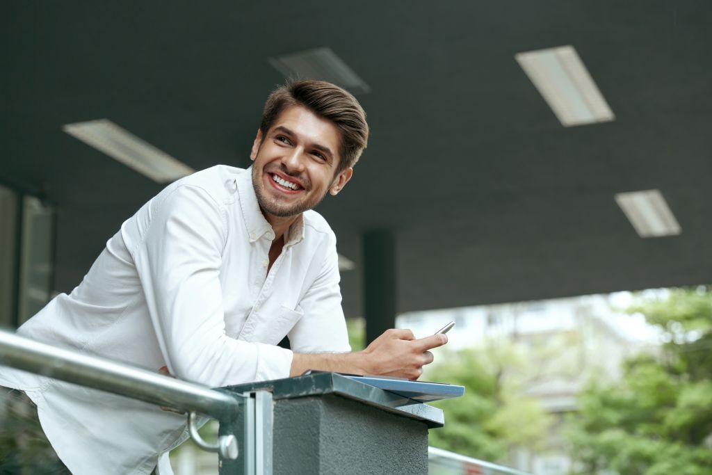 Smiling man in white long sleeves holding a smartphone while leaning against a balcony railing outdoors